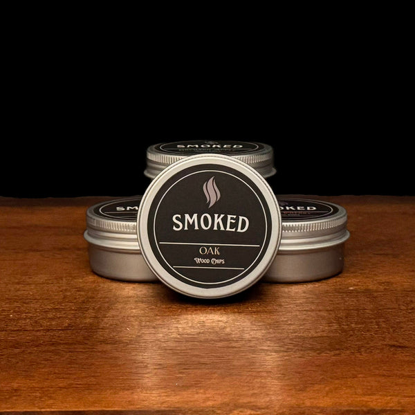 Four tins labeled 'Smoked' on a wooden surface with a black background. The front tin is Oak Flavored