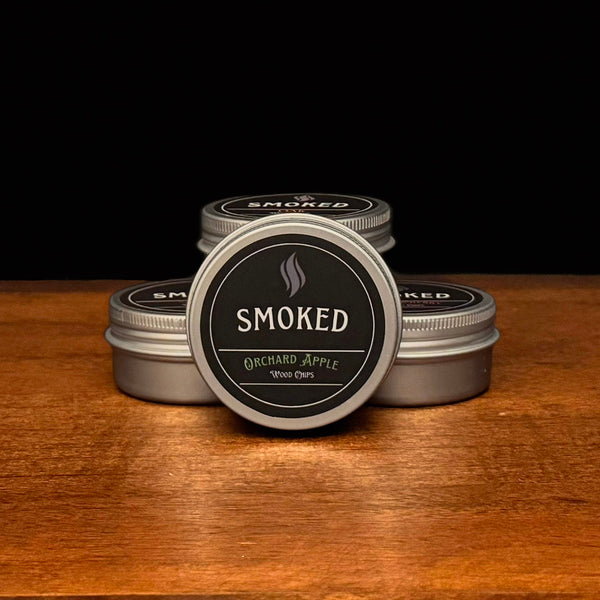 Four tins labeled 'Smoked' on a wooden surface with a black background. The front tin is Orchard Apple Flavored.