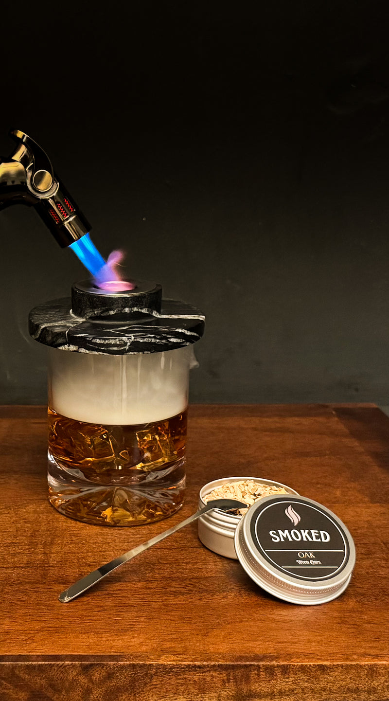 Glass with liquid being heated by a blowtorch on a wooden surface, with 'Smoked' branded items nearby.
