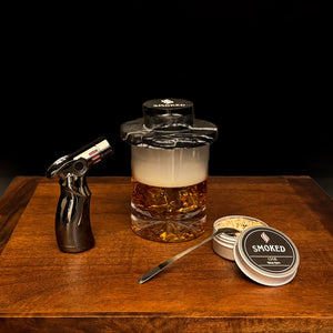 Glass of Smoked whiskey with a torch, spoon, and wood chip container on a wooden surface. The marble smoker is on top of the whiskey glass.