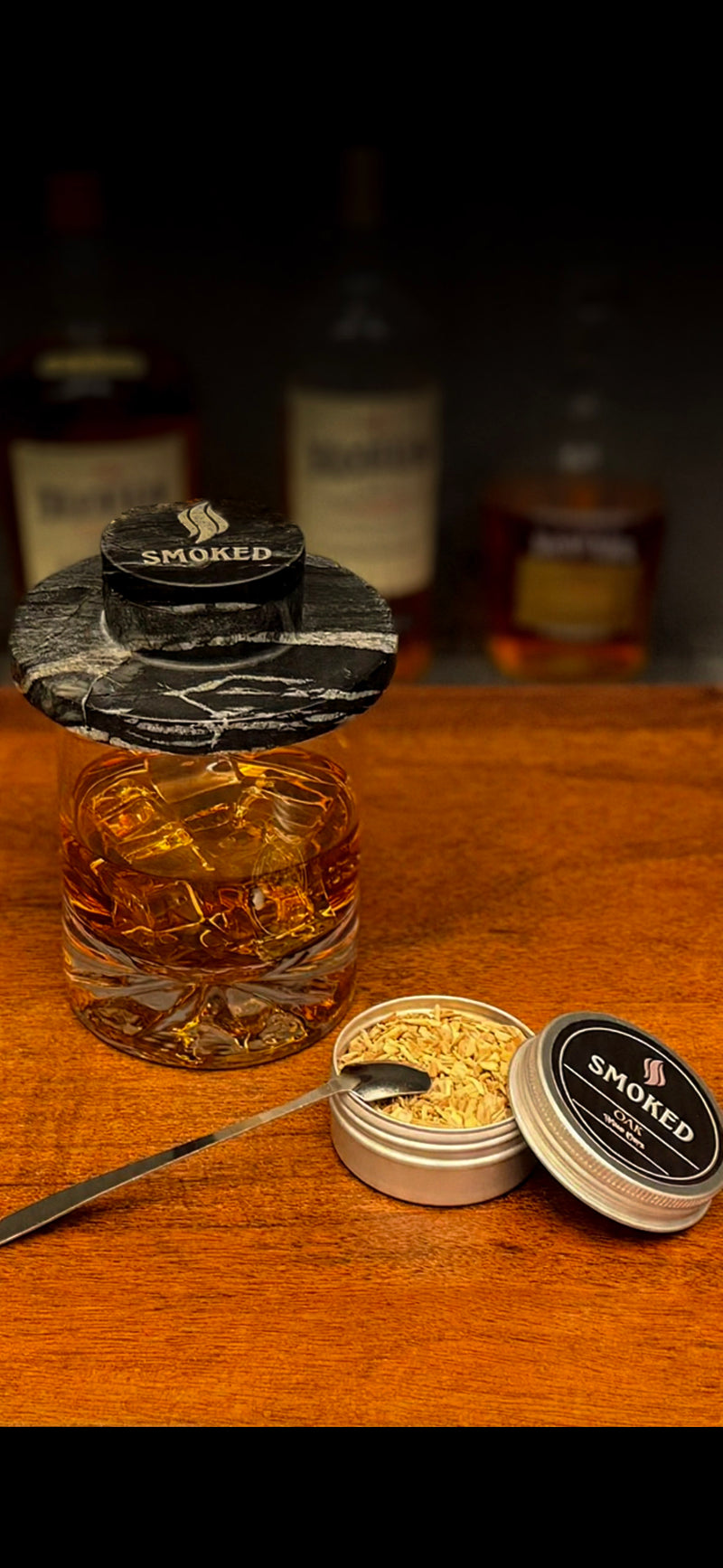 Glass of whiskey with ice cubes on a wooden surface, surrounded by bottles and smoking accessories.