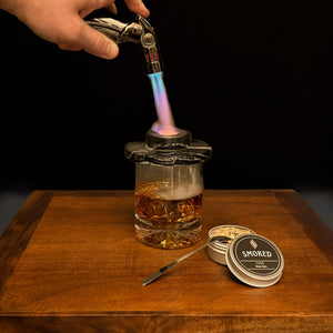 Hand using a butane lighter to light a glass of alcohol on a wooden surface with 'Smoked' product packaging.